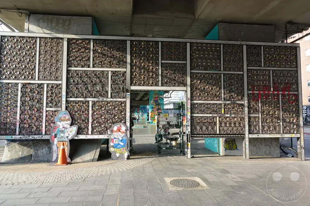 Eine eindrucksvolle Installation aus Computerplatinen, bedeckt große Metallflächen unter einer Brücke in Taipehs Zhongzheng District. Neben der Kunstinstallation stehen Pappaufsteller von Figuren, die di e urbane Atmosphäre des Ortes unterstreichen. Der Durchgang in den Hintergrund enthüllt einen belebten Skaterpark, der von bunter Straßenkunst geprägt ist und eine lebendige, jugendliche Stimmung vermittelt.