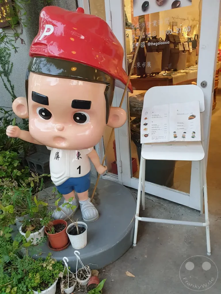 Eine auffällige, lebensgroße Cartoon-Figur mit roter Baskenmütze steht vor einem Geschäft im Huashan 1914 Creative Park in Taipeh, Taiwan. Neben der Figur ist ein weißer Ständer mit einer Speisekarte platziert, der auf das kulinarische Angebot oder die Ausrichtung des Geschäfts hinweist. Die Szene strahlt eine verspielte und einladende Atmosphäre aus, die typisch für kreative Kulturzentren ist.