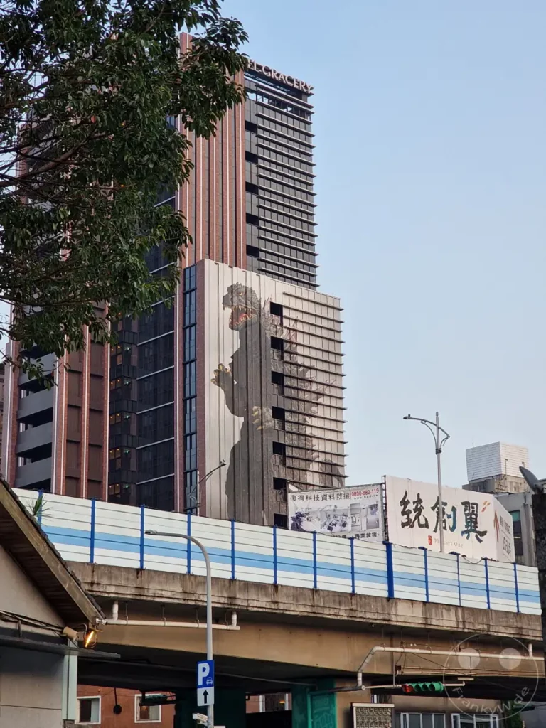 Ein riesiger Godzilla ziert die Fassade eines modernen Hochhauses in Taipeh, Taiwan. Über einer erhöhten Straße mit Verkehrsschildern erhebt sich die beeindruckende Architektur des Gebäudes, gekrönt von der Aufschrift "HOTEL GRACERY". Die Szene, eingefangen im klaren Tageslicht, vermittelt ein Gefühl von urbaner Dynamik und popkultureller Präsenz inmitten der kreativen Umgebung des Huashan 1914 Creative Parks.