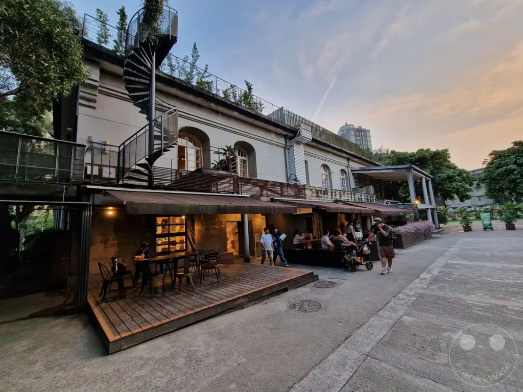 Der Huashan 1914 Creative Park in Taipeh, Taiwan, präsentiert sich am Abend mit seiner beeindruckenden historischen Architektur und einer belebten Außenanlage. Menschen genießen die entspannte Atmosphäre auf der Holzterrasse vor dem Gebäude. Die Szene wird von der warmen Abendsonne beleuchtet und strahlt eine friedliche Stimmung aus.
