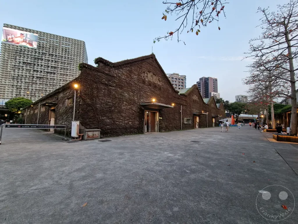 Historische Gebäude, die mit Pflanzen bewachsen sind, prägen den Huashan 1914 Creative Park in Taipeh, Taiwan. Moderne Architektur und belebte Wege mit sitzenden Menschen bilden einen Kontrast zum alten Baumbestand und den begrünten Fassaden. Die Szene strahlt eine ruhige Abendstimmung inmitten des städtischen Treibens aus.