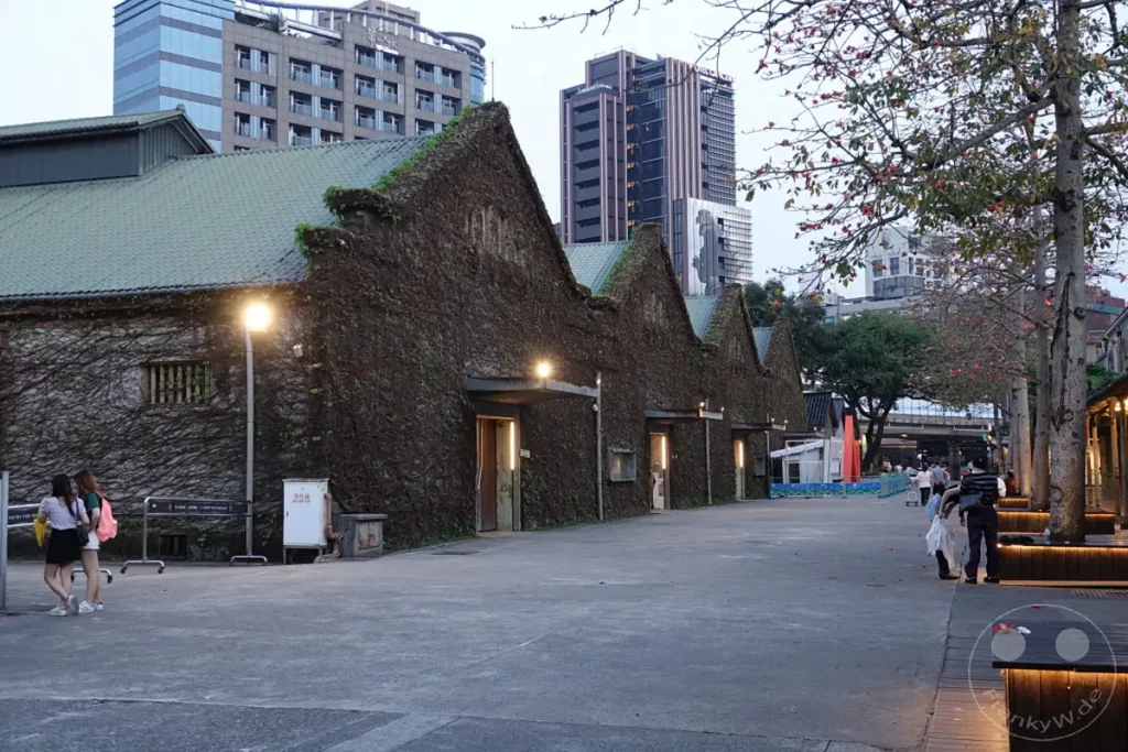 Historische Gebäude, die mit Efeu bedeckt sind, säumen einen weitläufigen gepflasterten Platz im Huashan 1914 Creative Park in Taipeh, Taiwan. Eingänge, die mit Lichtern beleuchtet sind, deuten auf Innenräume hin, während moderne Hochhäuser im Hintergrund aufragen. Die Szene vermittelt eine ruhige Atmosphäre, die Alt und Neu in der pulsierenden Stadt miteinander verbindet.