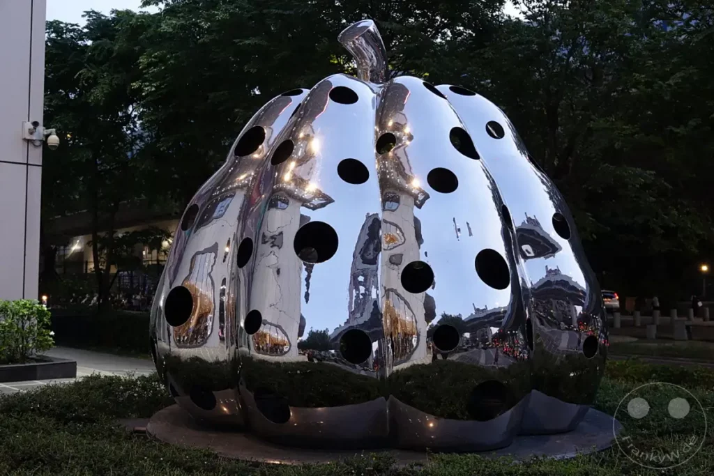 Eine glänzende Edelstahl-Kürbisskulptur mit vielen runden Aussparungen steht in einem städtischen Außenbereich in Taipeh, Taiwan. Die reflektierende Oberfläche fängt die umliegende Architektur und den bewölkten Himmel ein und erzeugt ein interessantes Spiel aus Licht und Schatten. Die Skulptur ist Teil des Zhongzheng Districts und verleiht dem öffentlichen Raum eine moderne künstlerische Note.
