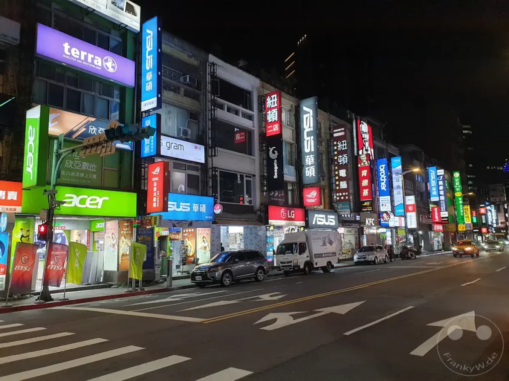 Nachts ist die Bade Road in Taipeh, Taiwan, als "PC-Meile" bekannt und beeindruckt durch die grellen Neonschilder von unzähligen Elektronikgeschäften. Geschäfte, die sich auf Computer und Technik spezialisiert haben, reihen sich aneinander und beleuchten die belebte Straße mit ihren bunten Lichtern. Die Szene vermittelt eine lebhafte und geschäftige Atmosphäre, typisch für eine städtische Einkaufsstraße, die bis spät in die Nacht geöffnet hat.