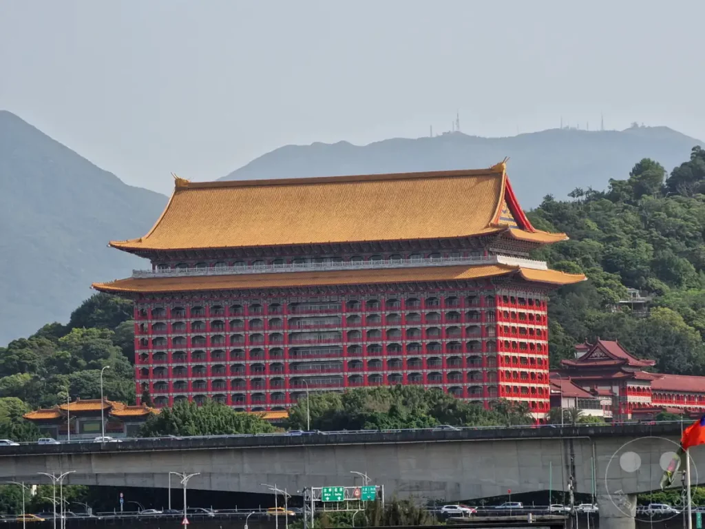 Das imposante Grand Hotel in Taipeh, Taiwan, dominiert mit seiner traditionellen roten Fassade und dem charakteristischen gelben Dach den Anblick. Im Hintergrund erheben sich bewaldete Hügel und Berge, während eine Autobahnbrücke mit Verkehr im Vordergrund die städtische Infrastruktur symbolisiert. Die Szene vermittelt einen Eindruck von kultureller Pracht inmitten einer bergigen Landschaft.