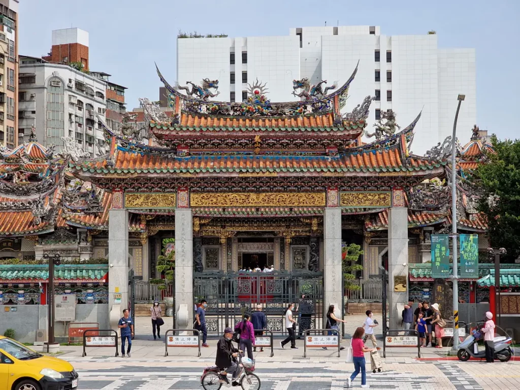 Der Mengjia Longshan Tempel in Taipeh, Taiwan, besticht durch seine prachtvolle traditionelle Architektur mit verzierten Dächern und Drachenfiguren. Der belebte Platz davor zeigt das alltägliche Leben in der Stadt, mit Menschen, die den Tempel besuchen oder vorbeigehen. Die Szene vermittelt eine lebendige Atmosphäre, in der historische Baukunst und modernes Stadtleben aufeinandertreffen.