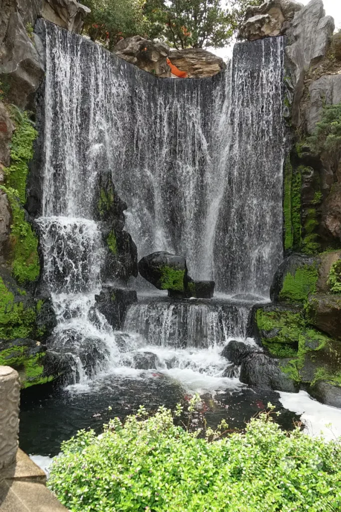 Ein beeindruckender Wasserfall stürzt in einen ruhigen Teich, umgeben von üppigem Grün und Felsen. Die Szenerie im Mengjia Longshan Tempel Park in Taipeh, Taiwan, strahlt eine friedliche und natürliche Atmosphäre aus. Koi-Karpfen sind im Wasser unter dem Wasserfall zu erkennen, was die Szene lebendig macht.
