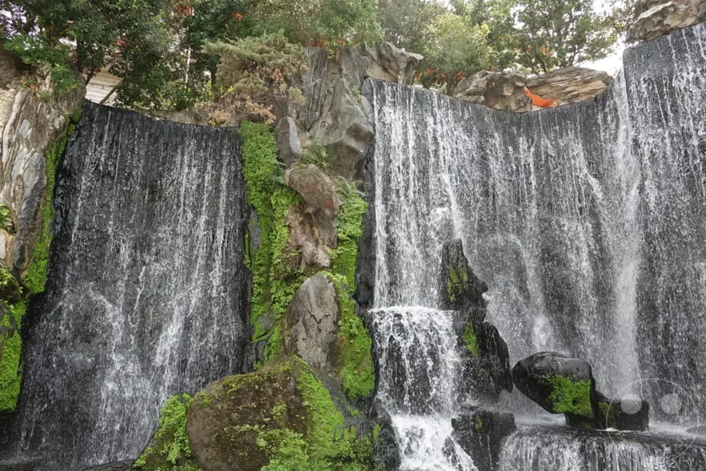 Ein beeindruckender künstlicher Wasserfall stürzt über moosbewachsene Felsen und eine steinerne Mauer herab. Das Wasser fällt in mehreren Kaskaden und schafft eine beruhigende und natürliche Atmosphäre inmitten der städtischen Umgebung des Mengjia Longshan Tempels in Taipeh, Taiwan. Die Szene ist von üppigem Grün und Felsformationen umgeben, die das Design des Tempelkomplexes ergänzen.