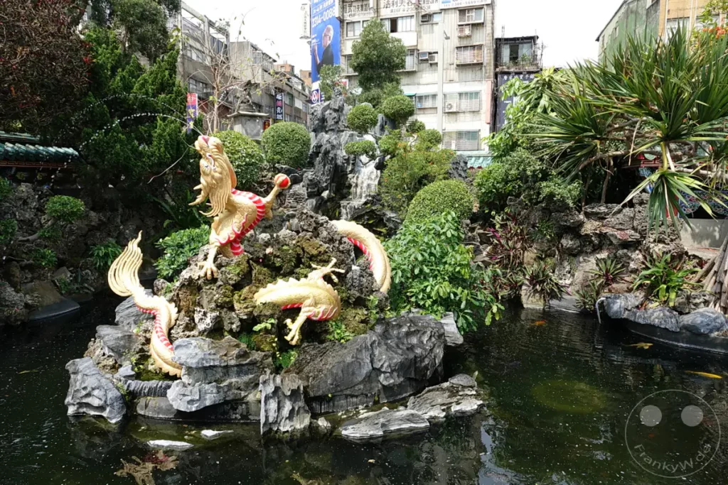 Goldene Drachenfiguren thronen auf einem felsigen Wasserfall, umgeben von üppiger Vegetation und einem Teich im Mengjia Longshan Tempel in Taipeh, Taiwan. Das lebhafte Arrangement symbolisiert traditionelle chinesische Kultur und schafft eine friedliche, spirituelle Atmosphäre. Koi-Karpfen schwimmen im dunklen Wasser, während im Hintergrund eine städtische Kulisse sichtbar ist.