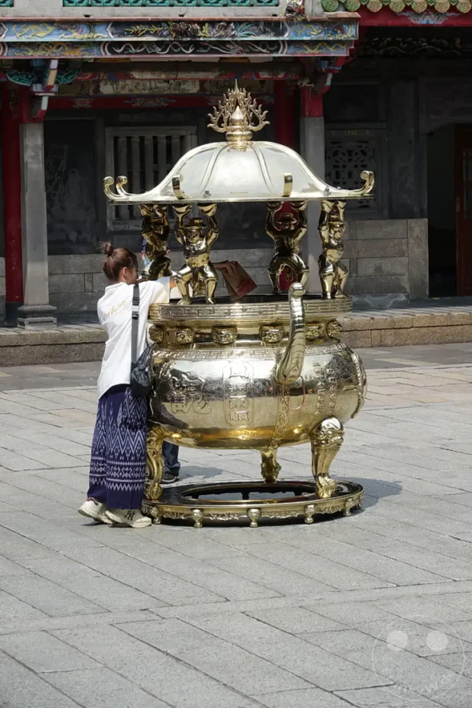 Eine Frau verharrt in Gedanken vor einem kunstvoll verzierten, goldenen Weihrauchgefäß im Mengjia Longshan Tempel in Taipeh, Taiwan. Die Sonne wirft lange Schatten auf den gepflasterten Hof, was eine friedliche und ehrfürchtige Atmosphäre schafft. Das detailreiche Gefäß dominiert den Vordergrund, während die traditionelle Tempelarchitektur im Hintergrund zu sehen ist.