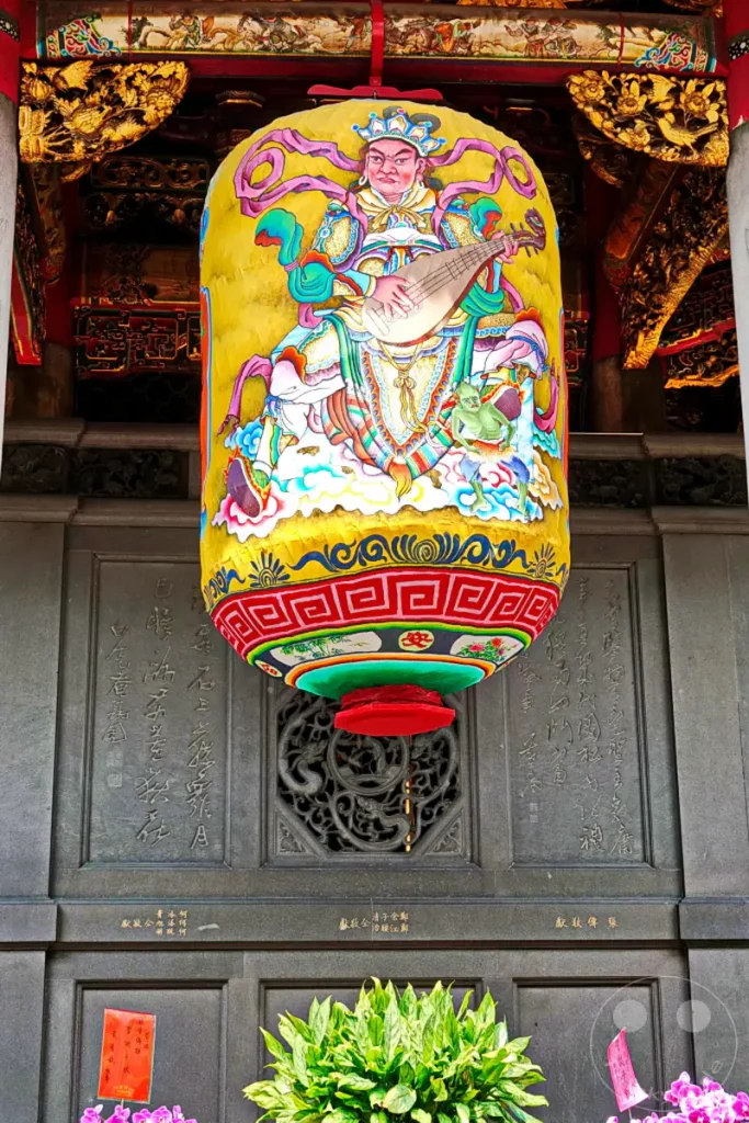 Eine große, gelbe Stofflaterne mit einer farbenfrohen Darstellung einer Gottheit im traditionellen Stil hängt vor einem detailreichen Tempelgebäude in Taipeh, Taiwan. Die aufwendige Verzierung der Laterne, mit einer spielenden Figur und Wolkenmotiven, sowie die architektonischen Elemente des Mengjia Longshan Tempels erzeugen eine feierliche und kulturell reiche Atmosphäre. Der Blick auf die Inschriften und das kunstvolle Gitterwerk am Gebäude unterstreicht die historische und spirituelle Bedeutung des Ortes.