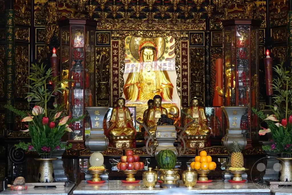 Im Mengjia Longshan Tempel in Taipeh, Taiwan, bildet eine prächtige goldene Buddha-Statue den Mittelpunkt eines reich verzierten Altars. Zahlreiche kleinere goldene Buddha-Figuren und Opfergaben wie Früchte schmücken die Szenerie, die von einer Atmosphäre tiefer Spiritualität und kultureller Bedeutung geprägt ist.
