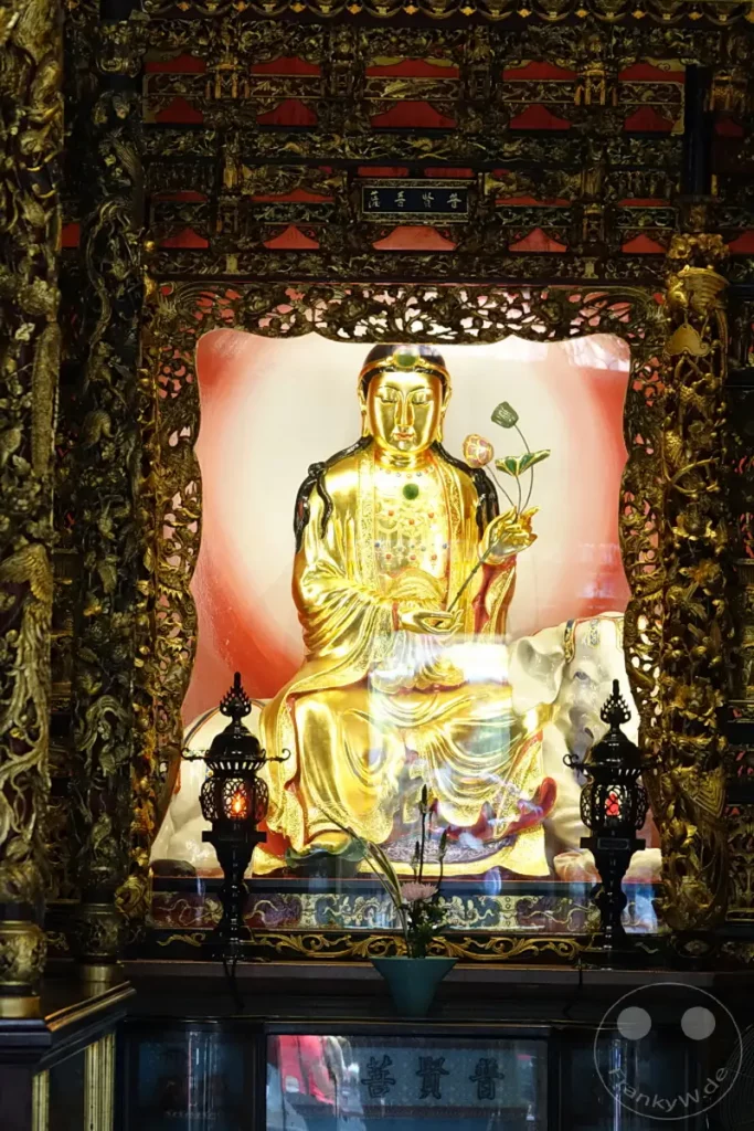 In einem reich verzierten goldenen Schrein in Taipeh, Taiwan, thront eine Buddha-Statue auf einem weißen Elefanten. Die Szene strahlt eine friedliche und ehrfürchtige Atmosphäre aus, unterstrichen durch die kunstvollen Verzierungen und die leuchtende Aura um die Gottheit im Mengjia Longshan Tempel. Kerzenlicht und ein Blumenarrangement beleben den Bereich vor dem Schrein und laden zum Verweilen ein.