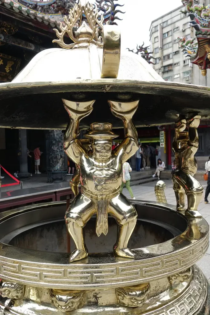 Eine detailreiche Messingfigur trägt den oberen Teil eines großen, verzierten Weihrauchfasses im Innenhof des Mengjia Longshan Tempels in Taipeh, Taiwan. Im Hintergrund sind Gebäude und andere Tempelstrukturen zu sehen, die dem Bild eine historische und kulturelle Atmosphäre verleihen. Die glänzende Oberfläche des Metalls und die komplexen Gravuren dominieren die Szene und unterstreichen die kunsthandwerkliche Qualität des Objekts.