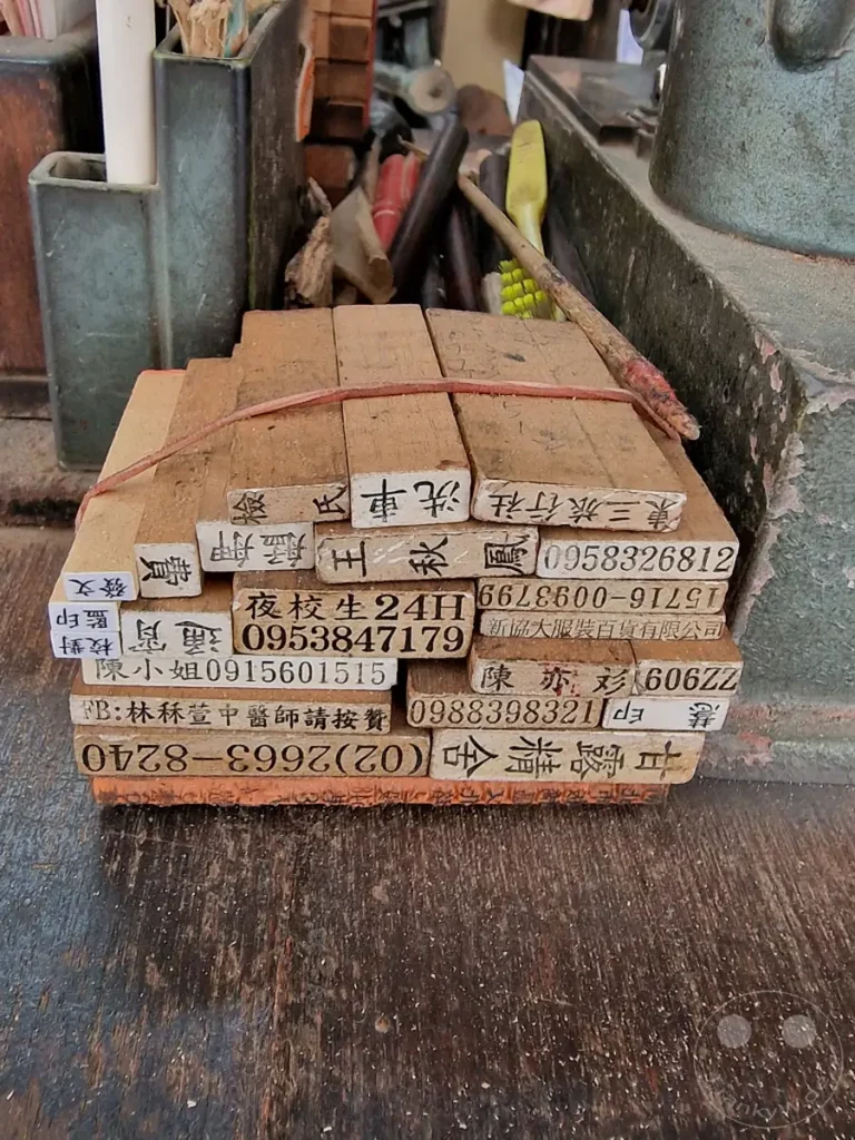 Eine Ansammlung alter Holzstempel mit chinesischen Schriftzeichen und Telefonnummern liegt auf einer Holztischplatte in Taipeh. Die Stempel sind mit einem roten Gummiband zusammengebunden und vermitteln eine nostalgische Atmosphäre, die an die Handwerkskunst vergangener Zeiten erinnert. Staub und Abnutzungsspuren auf den Stempeln zeugen von ihrer intensiven Nutzung und ihrer Geschichte als Werkzeuge eines Stempelmachers.