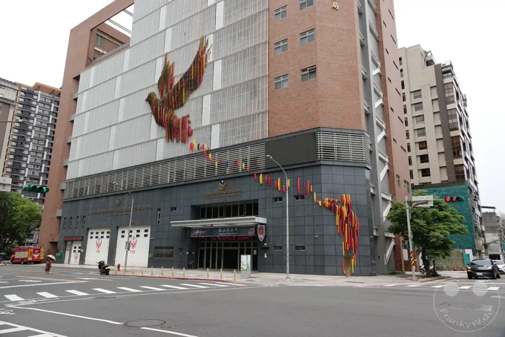 Das Wanhua Taipei City Government Police Bureau in Taiwan ist mit einem markanten Kunstwerk einer Taube geschmückt, das sich über die Fassade erstreckt. Rote und gelbe Elemente winden sich die Seiten des Gebäudes empor und bilden eine dynamische künstlerische Darstellung. Im Vordergrund des Bildes sind eine Straße und Fußgängerüberwege zu sehen, die die städtische Umgebung des Polizeigebäudes hervorheben.