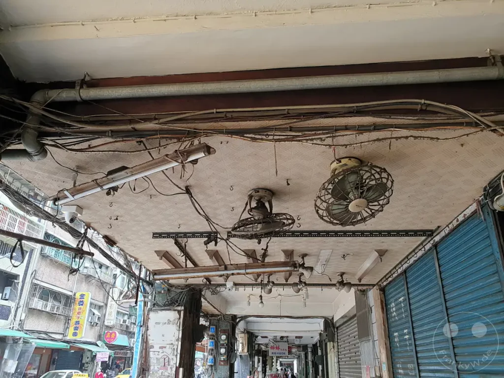 Diese Aufnahme aus Taipeh, Taiwan, zeigt einen überladenen Deckenbereich mit zahlreichen Kabeln, Rohren und Lüftern zur Kühlung. Die chaotische Installation deutet auf eine geschäftige städtische Umgebung hin, in der Funktionalität über Ästhetik gestellt wird. Die Szene vermittelt den Eindruck von Hitze und dem Bedürfnis nach effektiver Lüftung in der tropischen Stadt.