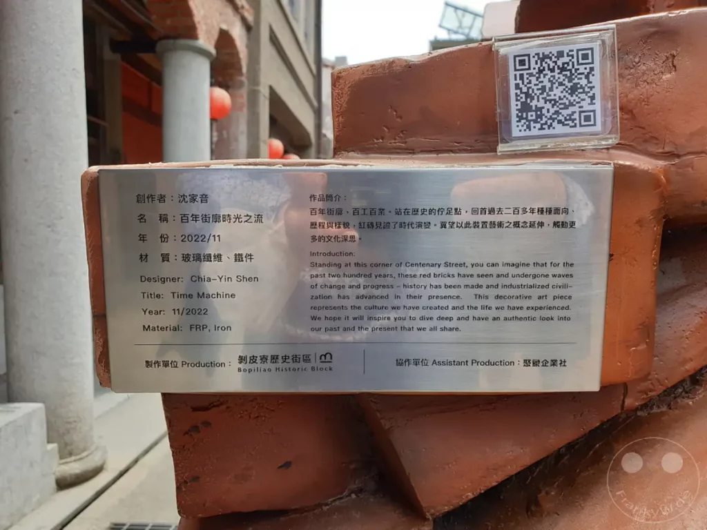 Eine Informationstafel vor einer rötlichen Skulptur im Bopiliao Historical Block in Taipeh, Taiwan, beschreibt das Kunstwerk "Time Machine". Die Skulptur soll die lange Geschichte des Ortes und die Entwicklung der Zivilisation widerspiegeln. Die Tafel enthält sowohl chinesische als auch englische Texte, die über die Gestaltung und die Bedeutung des Kunstwerks informieren.