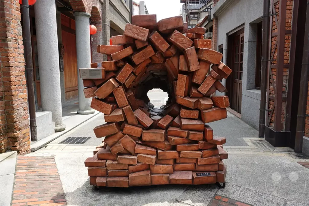 Eine künstlerische Skulptur aus roten Ziegelsteinen mit einem kreisförmigen Durchgang steht auf einem gepflasterten Weg im historischen Bopiliao Historical Block in Taipeh, Taiwan. Die Ziegel sind unregelmäßig angeordnet und schaffen eine organische, architektonische Form, die zum Durchschauen einlädt. Die Szene vermittelt einen Eindruck von historischem Charme und kreativer Nutzung traditioneller Materialien in einer städtischen Umgebung.