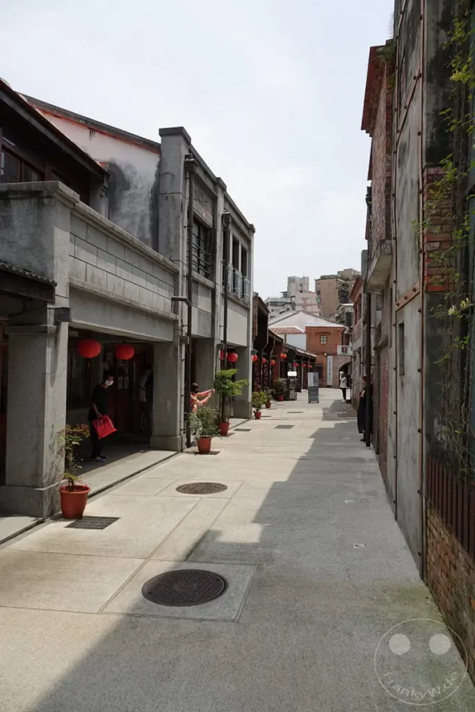Eine schmale Gasse mit historischen Gebäuden im Bopiliao Historical Block in Taipeh, Taiwan, lädt zum Spaziergang ein. Rote Laternen und Pflanzen schmücken die Fassaden und verleihen der Szene eine kulturelle Note. Die Architektur und die ruhige Atmosphäre lassen vergangene Zeiten lebendig werden.