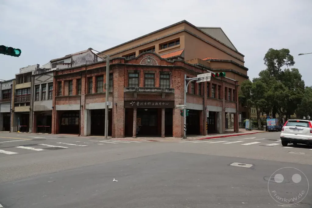 Ein Gebäudeensemble mit traditioneller Architektur im Bopiliao Historical Block in Taipeh, Taiwan, steht an einer belebten Kreuzung. Die Backsteinbauten und die moderne angrenzende Struktur bilden einen Kontrast vor einem bewölkten Himmel. Eine weiße SUV und ein Lieferwagen sind auf der Straße zu sehen, was die alltägliche Atmosphäre der historischen Stätte unterstreicht.