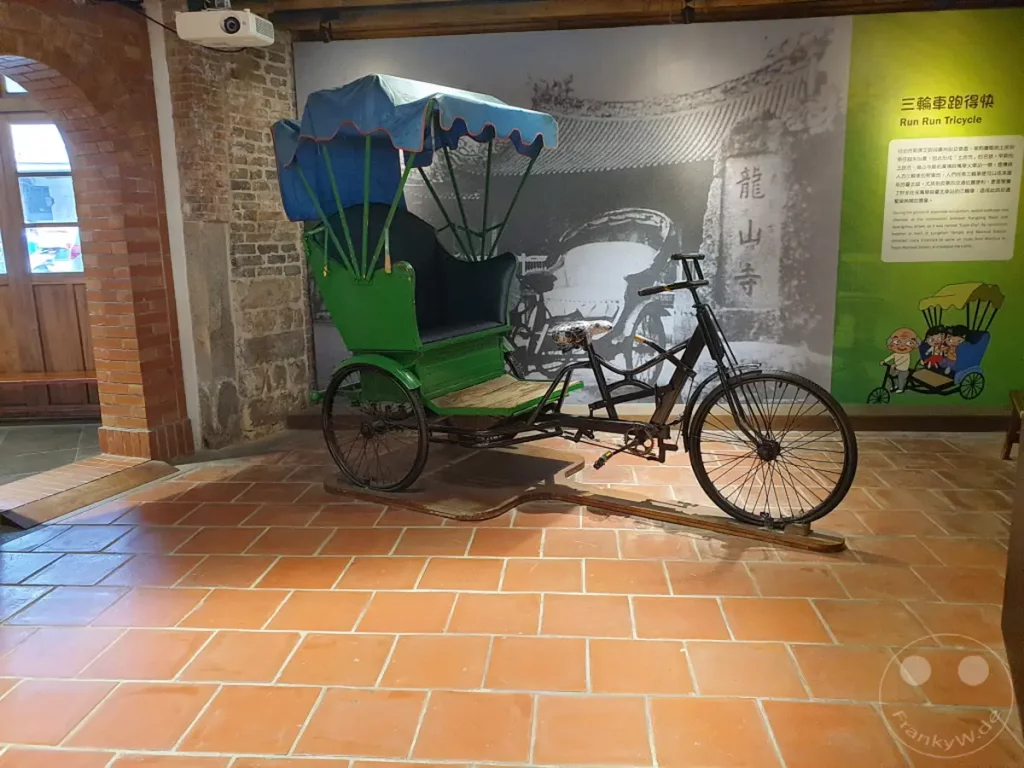 In Taipeh, im Bopiliao Historical Block, steht eine restaurierte grüne Rikscha als Ausstellungsstück. Die Rikscha ist vor einer Wand mit historischen Schwarz-Weiß-Fotos und einem farbigen Bild einer Rikscha mit Passagieren positioniert. Die Szenerie vermittelt einen nostalgischen Einblick in die Transportgeschichte Taiwans.