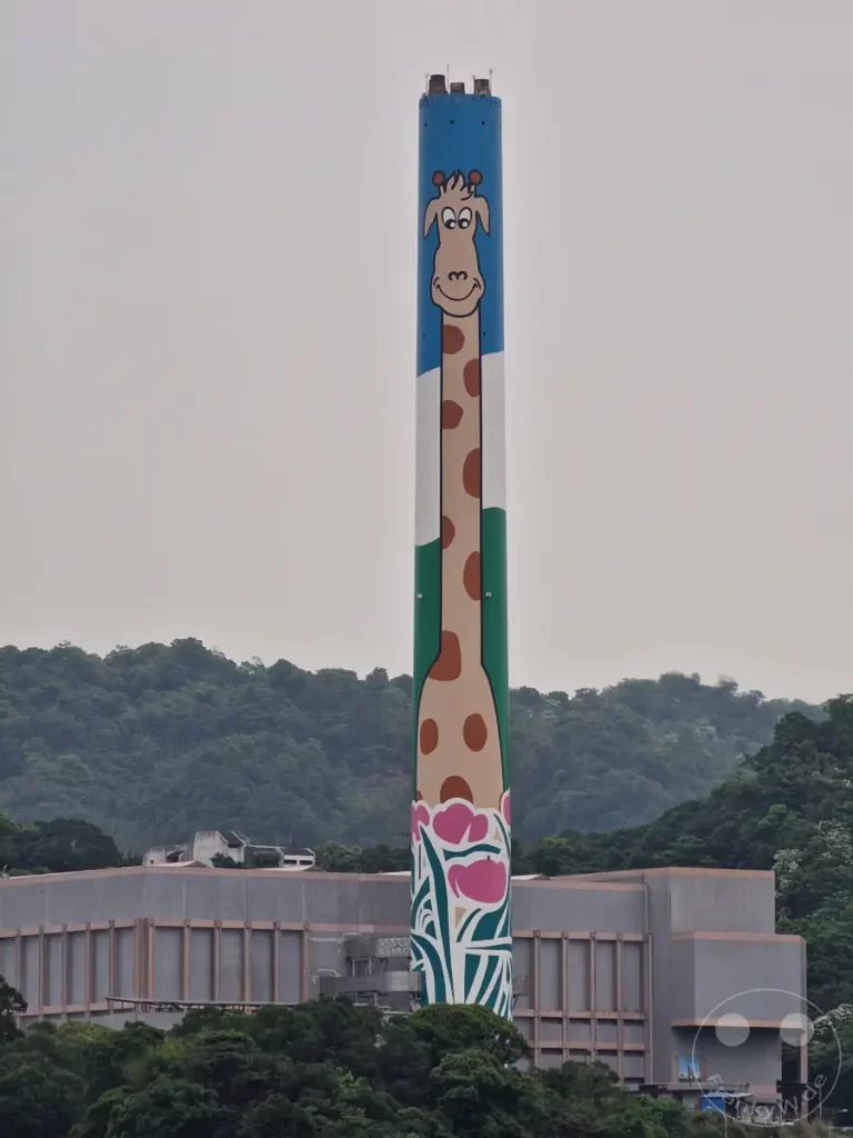 Ein markanter Schornstein, bemalt mit einer fröhlichen Giraffe, steht in Taipeh, Taiwan, hinter einer Industrieanlage. Die bunte Gestaltung des Turms, die auch Blumenmotive am Sockel aufweist, bildet einen unerwarteten Kontrast zur sonst eher grauen Umgebung. Diese auffällige Kunstinstallation am Rande des Taipei Zoos verleiht der Kulisse eine spielerische und charmante Note.