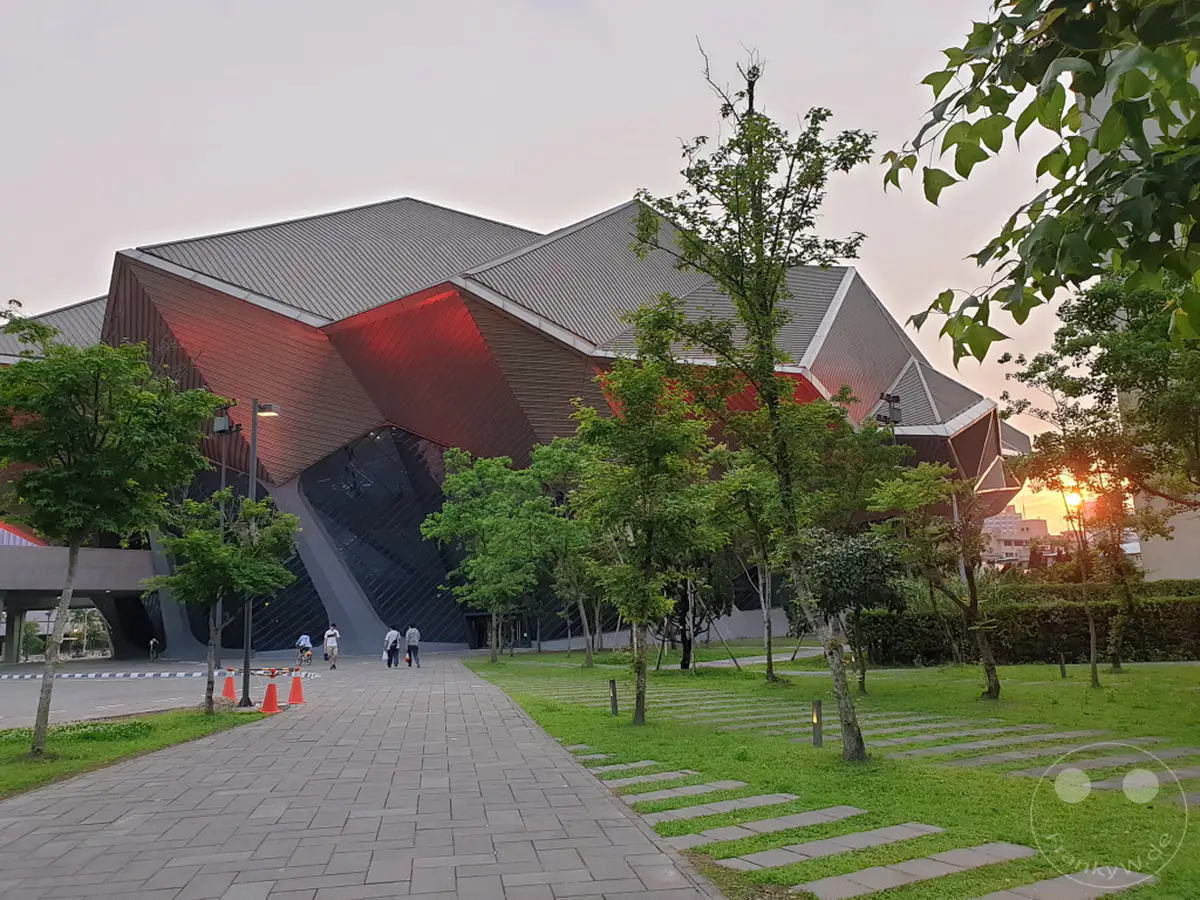 Das Taipei Music Center in Taiwan präsentiert sich in moderner, kantiger Architektur mit auffälliger roter Beleuchtung. Vor dem Gebäude erstreckt sich ein gepflasterter Weg durch eine grüne Parkanlage mit Bäumen, während im Hintergrund die Sonne untergeht und ein warmes Licht wirft. Die Szene vermittelt eine ruhige, aber architektonisch beeindruckende Abendstimmung in Taipeh.