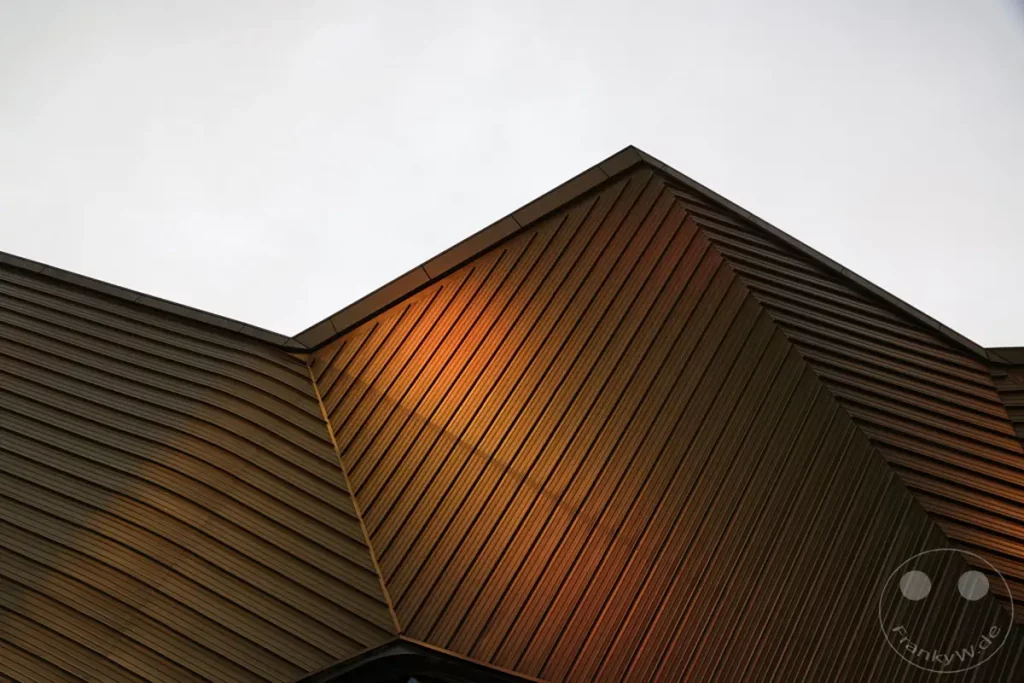 Die moderne Architektur des Taipei Music Center in Taipeh, Taiwan, präsentiert sich mit einer markanten, lamellenartigen Fassade. Sonnenlicht streift über die geneigten Elemente und erzeugt ein Spiel aus Licht und Schatten, das die geometrische Form des Gebäudes unterstreicht. Die Aufnahme fängt die beeindruckende Struktur und die atmosphärische Beleuchtung des kulturellen Zentrums ein.