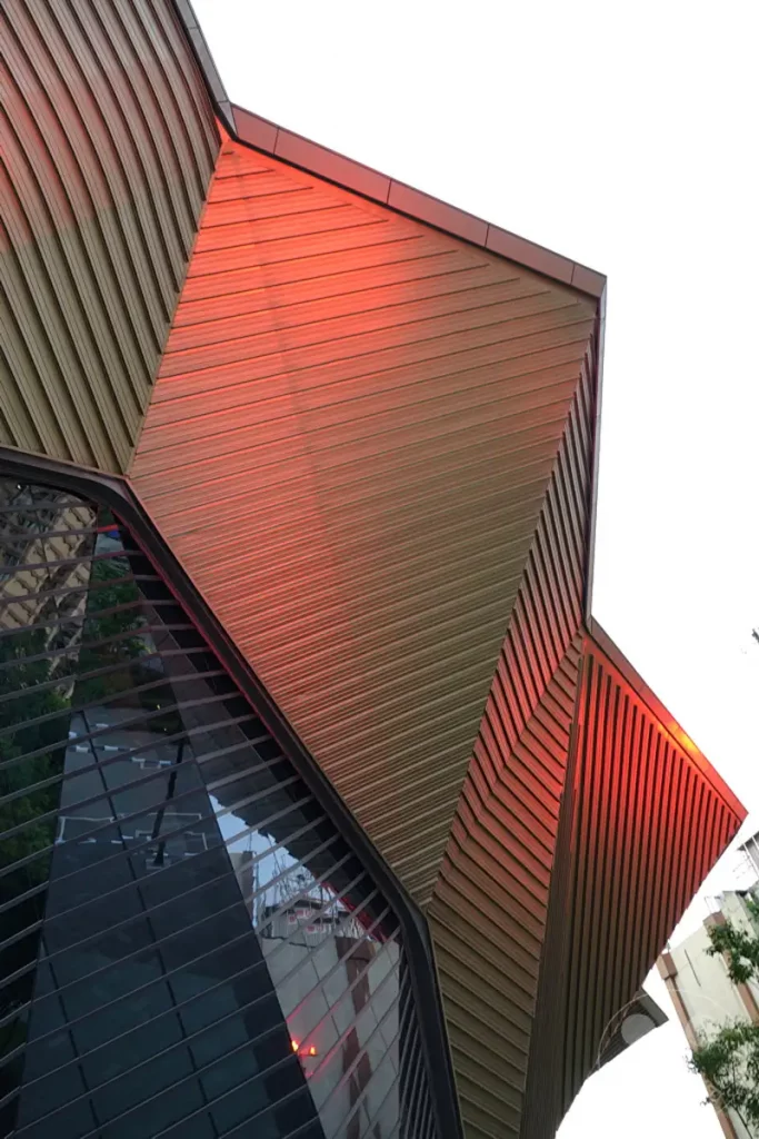 Die moderne Architektur des Taipei Music Center in Taipeh, Taiwan, wird durch das warme Licht der Abendsonne in Rottönen beleuchtet. Geometrische Formen und Glasflächen prägen die dynamische Fassade des Gebäudes. Dieses Zusammenspiel von Licht und Struktur erzeugt eine beeindruckende visuelle Wirkung in der urbanen Landschaft Taipehs.