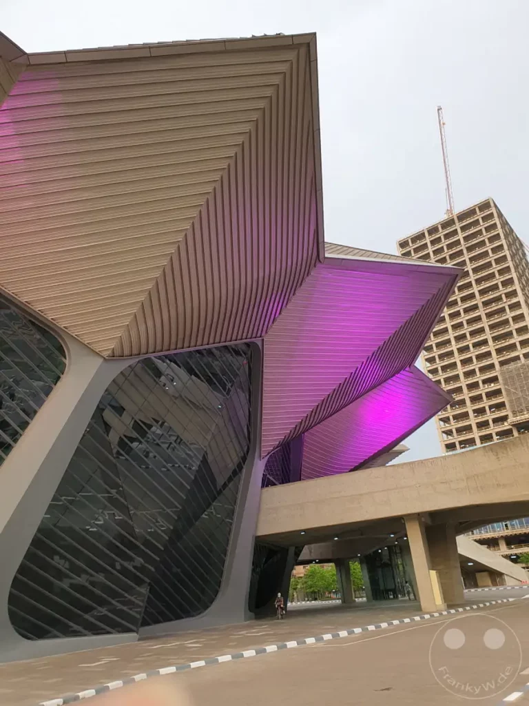 Die futuristische Architektur des Taipei Music Centers in Taipeh, Taiwan, erstrahlt in faszinierendem violettem Licht. Die geschwungenen Linien und die modernen Materialien des Gebäudes bilden einen starken Kontrast zu dem dahinterliegenden, klassischeren Hochhaus. Die Szene vermittelt eine Atmosphäre von urbaner Dynamik und künstlerischem Ausdruck.