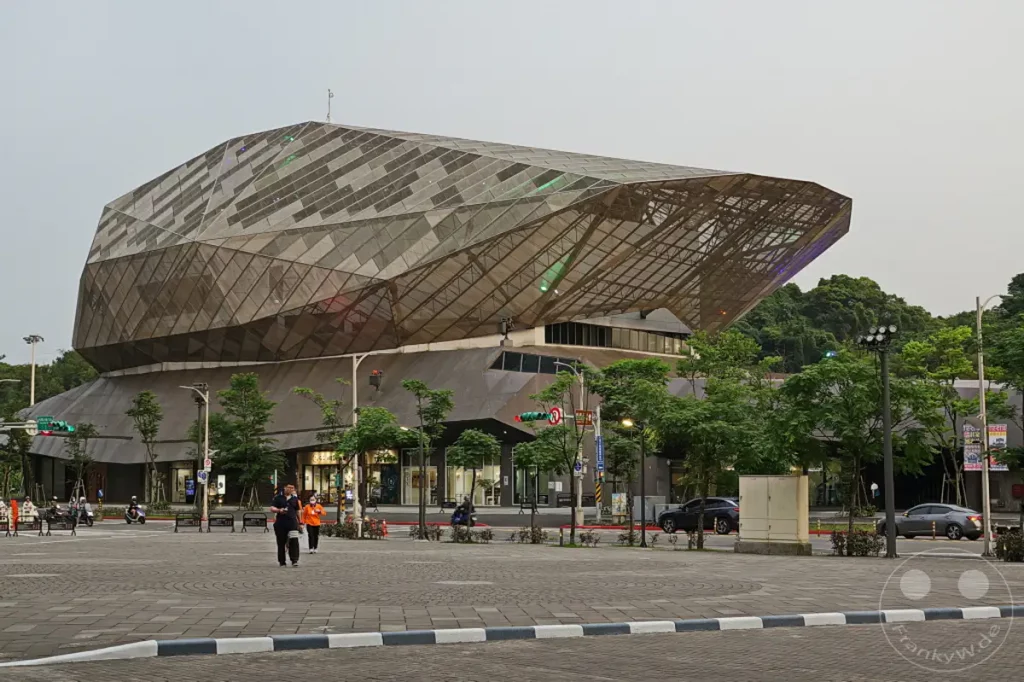 Ein markantes, futuristisch anmutendes Gebäude mit einer komplexen Glas- und Metallfassade prägt die Szenerie in Taipeh, Taiwan. Mehrere Bäume säumen den weitläufigen Platz vor dem Gebäude, auf dem sich einige Passanten und Fahrzeuge bewegen. Die Atmosphäre ist ruhig und leicht dämmrig, was der modernen Architektur eine besondere Note verleiht.