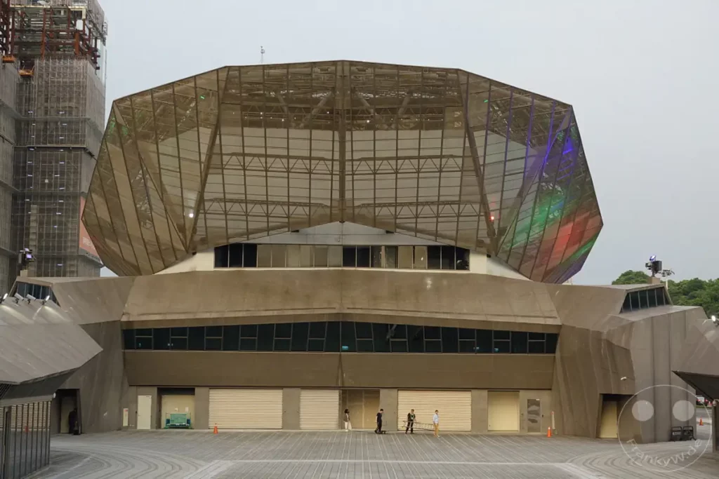 Die futuristische Architektur des Taipei Music Center Creative Hub dominiert die Szene unter einem bewölkten Himmel in Taipeh, Taiwan. Schichtweise gegliederte Betonelemente und eine markante, facettierte Glasstruktur prägen das Erscheinungsbild. Die Außensicht vermittelt einen Eindruck von moderner Industriekultur und städtischer Dynamik.