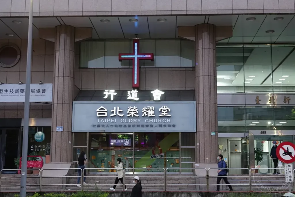 Die Taipei Glory Church in Taiwan ist ein imposantes Gebäude mit einem leuchtenden Kreuz über dem Eingang. Mehrere Menschen gehen an der Fassade vorbei, darunter einige mit Mundschutz, was auf die aktuelle Zeit hindeutet. Die Atmosphäre ist städtisch und leicht grau, was den Fokus auf die Architektur und das religiöse Symbol lenkt.