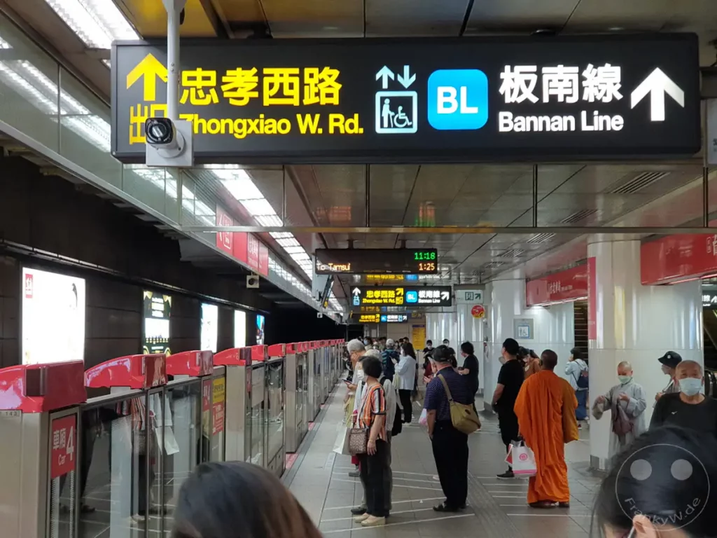 Ein belebter Bahnsteig der Metro in Taipeh, Taiwan. Fahrgäste warten auf die Bannan Line, während informative Schilder die Richtungen und Linien anzeigen. Die Atmosphäre ist geschäftig und urban, typisch für ein modernes U-Bahn-System.