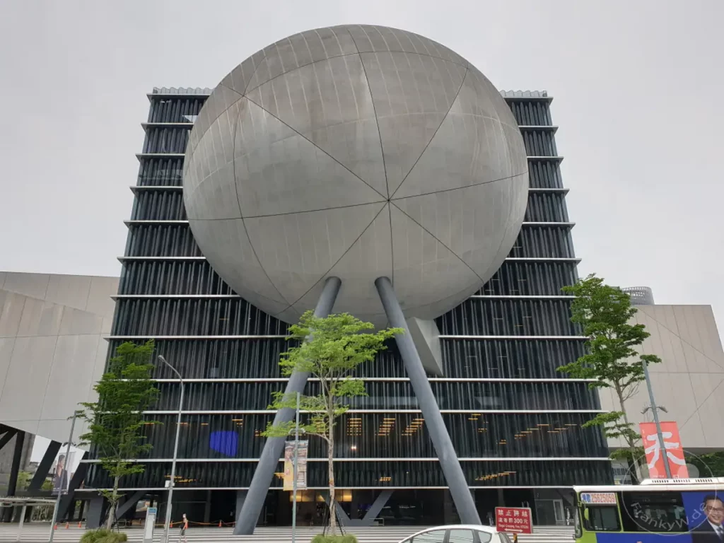 Das Taipei Performing Arts Center in Taipeh, Taiwan, dominiert mit seiner futuristischen Architektur und der riesigen kugelförmigen Struktur den Himmel. Die Fassade des Gebäudes ist mit vertikalen Lamellen versehen, die ihm ein dynamisches Aussehen verleihen. Grünes Laub und eine vorbeifahrende Buslinie schaffen eine lebendige städtische Atmosphäre vor dem imposanten Bauwerk.
