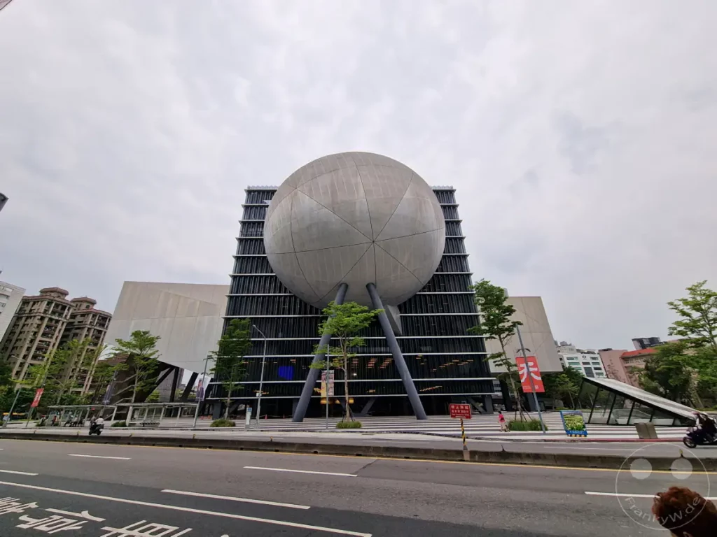 Das Taipei Performing Arts Center in Taipeh, Taiwan, präsentiert sich mit seiner markanten kugelförmigen Struktur und der modernen, geometrischen Fassade. Die Aufnahme fängt die einzigartige Architektur des Gebäudes im städtischen Umfeld ein. Ein bewölkter Himmel verleiht der Szene eine gedämpfte, aber beeindruckende Atmosphäre.