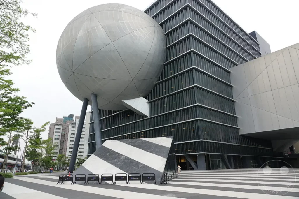 Das Taipei Performing Arts Center in Taipeh, Taiwan, präsentiert sich mit seiner futuristischen Architektur, die eine riesige Kugel auf einem modernen Gebäude mit vertikalen Lamellen vereint. Die kontrastreiche Gestaltung des Außenbereichs mit schwarz-weißen Streifen und einem schrägen, geometrischen Element schafft eine dynamische und künstlerische Atmosphäre. Die Szene fängt die avantgardistische Ästhetik dieses kulturellen Wahrzeichens ein und lädt zum Betrachten der einzigartigen Formen ein.
