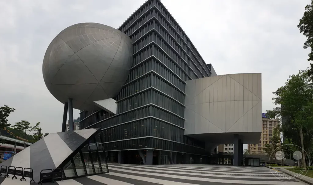 Das Taipei Performing Arts Center in Taipeh, Taiwan, präsentiert eine avantgardistische Architektur mit einer markanten Kugelform, die auf einem modernen Gebäudekomplex ruht. Klare Linien und geometrische Formen dominieren die Fassade und schaffen eine beeindruckende visuelle Präsenz. Die Szene vermittelt eine Atmosphäre von Innovation und künstlerischem Ausdruck in urbaner Umgebung.