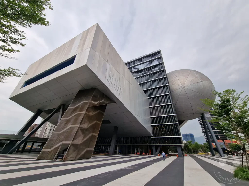 Das Taipei Performing Arts Center in Taipeh, Taiwan, präsentiert sich mit seiner avantgardistischen Architektur, die aus geometrischen Formen und schwebenden Elementen besteht. Die Fassade des Gebäudes, kombiniert mit der markanten kugelförmigen Struktur, schafft eine futuristische und beeindruckende Kulisse. Der weite Platz mit seinem schwarz-weiß gestreiften Muster und der leicht bewölkte Himmel verleihen der Szene eine ruhige und doch dynamische Atmosphäre.