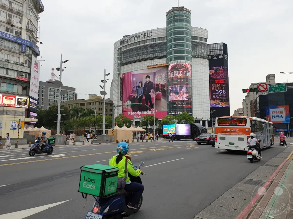 In Taipeh, Taiwan, pulsiert das Leben in der Nähe des Xinmen-Viertels, wo moderne Architektur auf geschäftiges Treiben trifft. Eine lebhafte Straßenszene entfaltet sich vor einem imposanten Einkaufszentrum, während der Verkehr durch die Stadt fließt. Fußgänger und Fahrer teilen sich den Raum und tragen zur dynamischen Atmosphäre bei.
