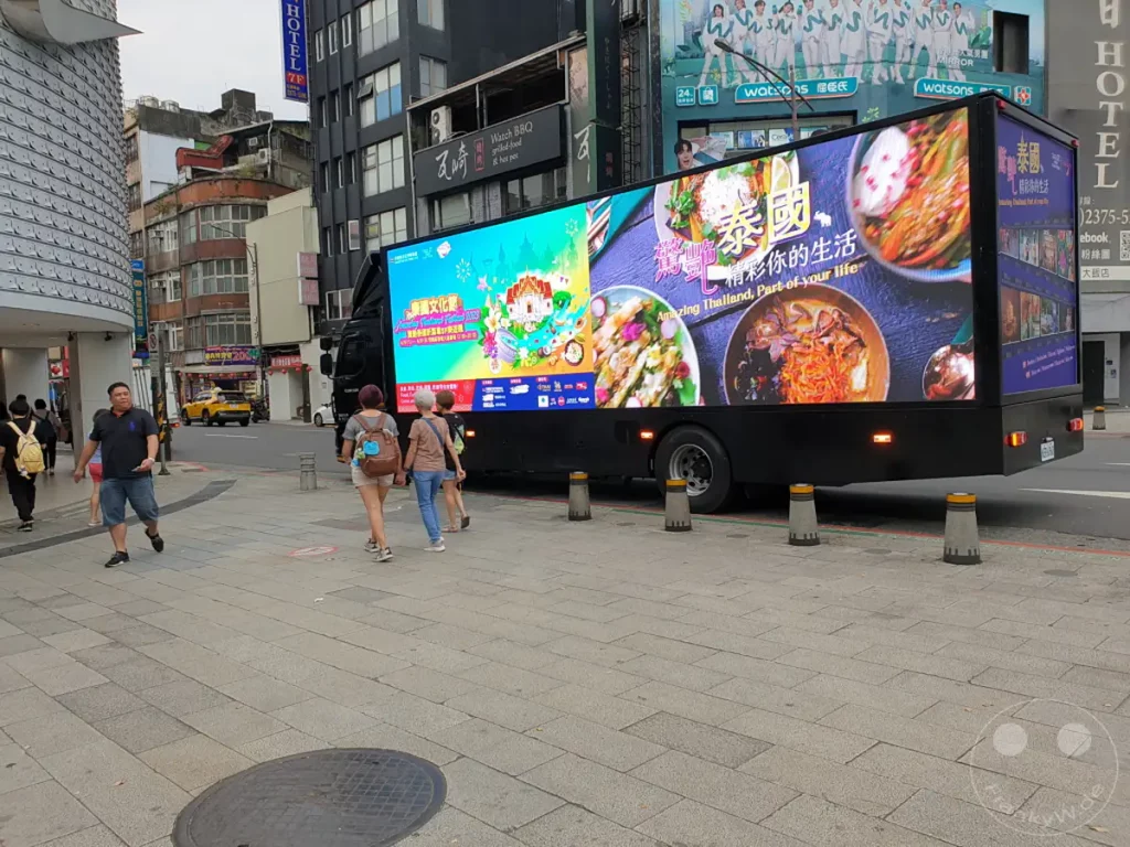 Ein leuchtender Werbe-LKW mit Bildern von thailändischem Essen und Schriftzügen steht in Taipeh, Taiwan, in der Nähe des belebten Viertels Xinmen. Mehrere Personen gehen auf dem Gehweg an dem Fahrzeug vorbei, während im Hintergrund städtische Gebäude und eine andere Werbung sichtbar sind. Die Szene vermittelt eine typische urbane Atmosphäre mit Fokus auf die mobile Werbeplattform.