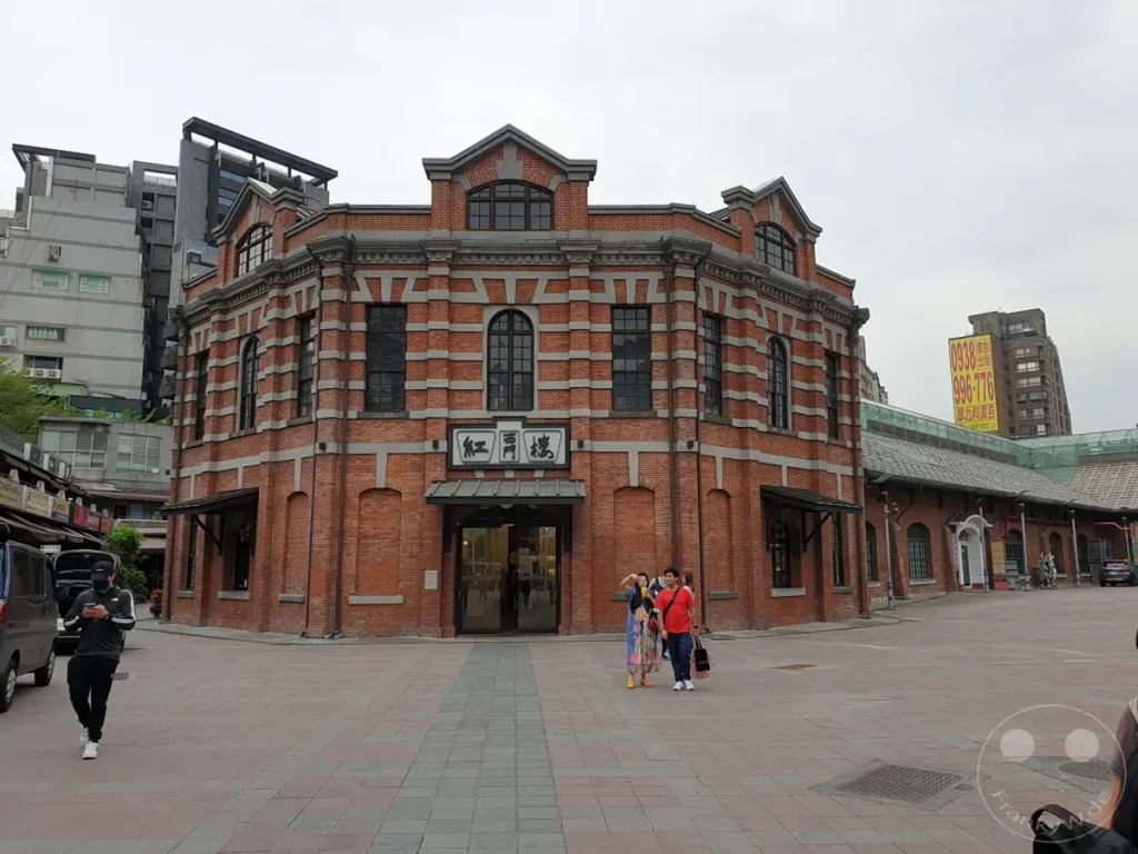 Das "The Red House" in Taipehs Xinmen-Viertel beeindruckt mit seiner roten Backsteinarchitektur im Kolonialstil. Menschen spazieren über den belebten Platz vor dem historischen Gebäude, das von modernen Bauten umgeben ist. Eine lebendige Atmosphäre entsteht durch die Mischung aus Tradition und urbanem Leben.