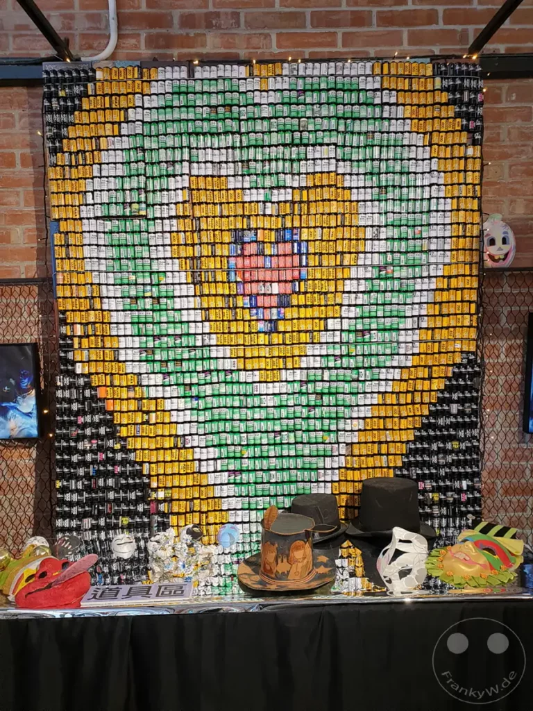 Eine beeindruckende Wandinstallation aus zahlreichen Filmdosen bildet ein Mosaik-Muster im Stil eines Arcade-Spiels, das im Bereich der Ausrüstung beim "The Red House" in Taipehs Xinmen Distrikt aufgestellt ist. Davor stehen mehrere Masken und Hüte, die zu einer theatralischen oder festlichen Atmosphäre beitragen. Die Szenerie wirkt bunt, kreativ und einladend, typisch für die lebhafte Umgebung von Xinmen.
