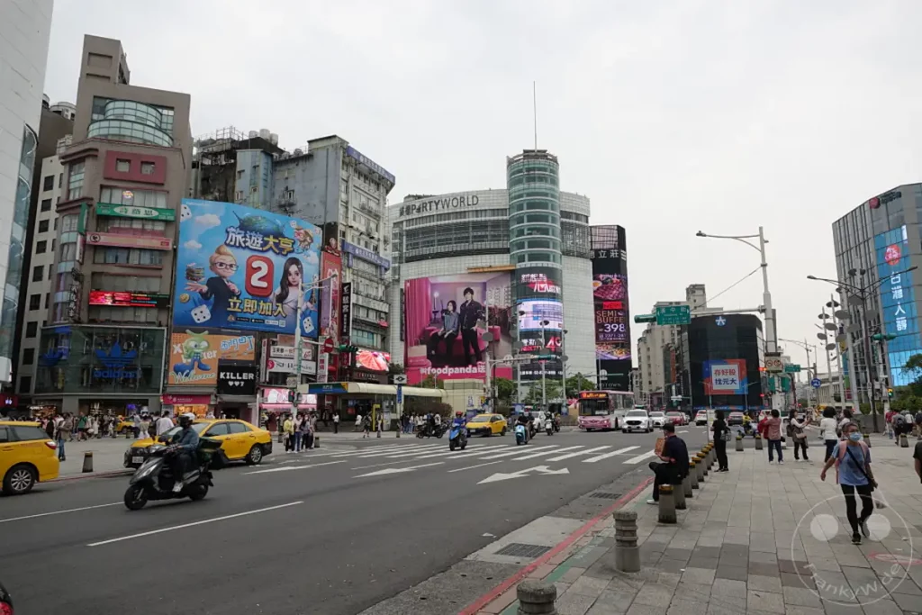Eine belebte Straße in Ximending, Taipeh, Taiwan, ist mit Geschäften, Restaurants und großen Werbetafeln geschmückt. Fußgänger und Fahrzeuge bevölkern die Szene, was die pulsierende Atmosphäre des beliebten Viertels widerspiegelt. Die Architektur ist eine Mischung aus modernen und älteren Gebäuden, die den einzigartigen Charakter der Gegend ausmachen.