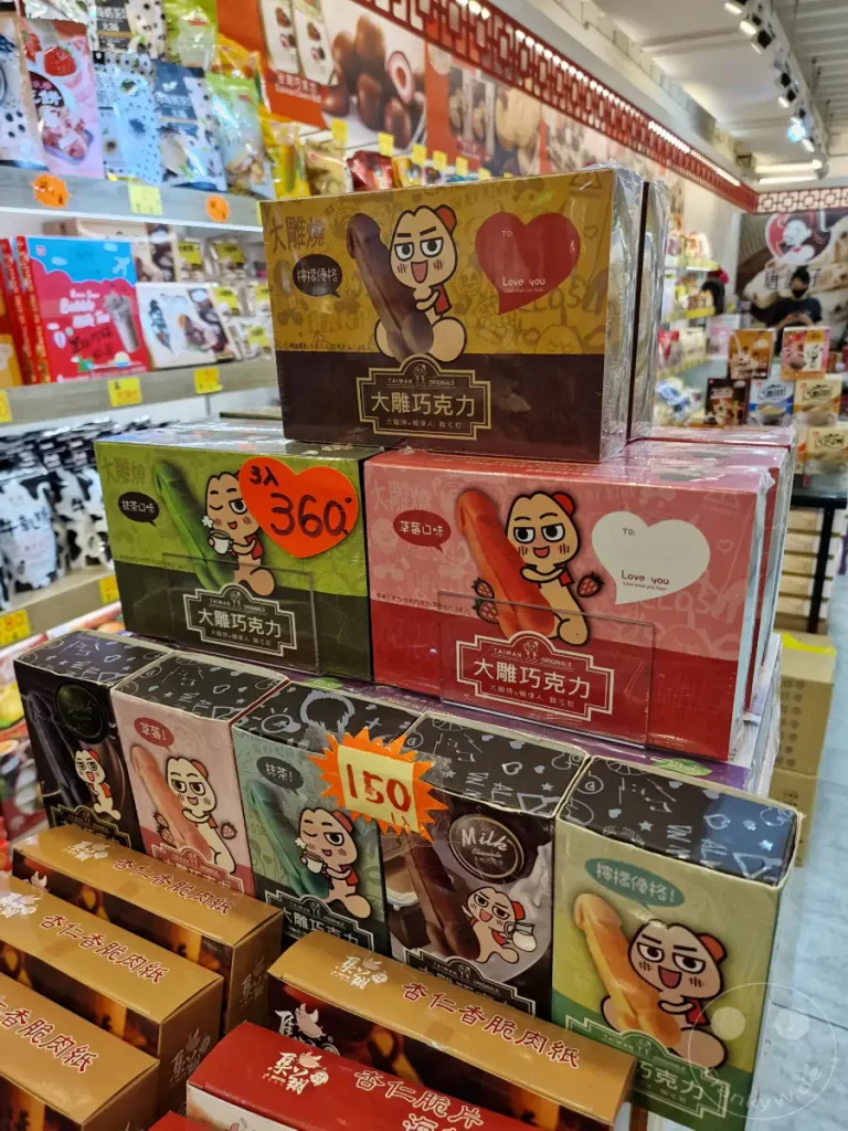 Eine farbenfrohe Auswahl an 'Cock Sweets' wird in einem Geschäft in Taiwan präsentiert. Die Verpackungen zeigen humorvolle Illustrationen und asiatische Schriftzeichen, was eine verspielte und originelle Atmosphäre schafft. Verschiedene Geschmacksrichtungen und Angebote sind zu sehen, die zum Kauf einladen.