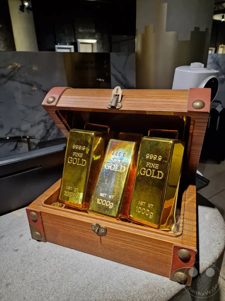 Drei glänzende Goldbarren mit der Aufschrift "999.9 FINE GOLD 1000g" liegen in einer hölzernen Schatzkiste. Das Setting in Taipeh, im Hotel Hua Shan Din, vermittelt ein Gefühl von Reichtum und Exklusivität. Die Darstellung ruft Assoziationen von Sicherheit, Wertanlage und der Faszination für Edelmetalle hervor.