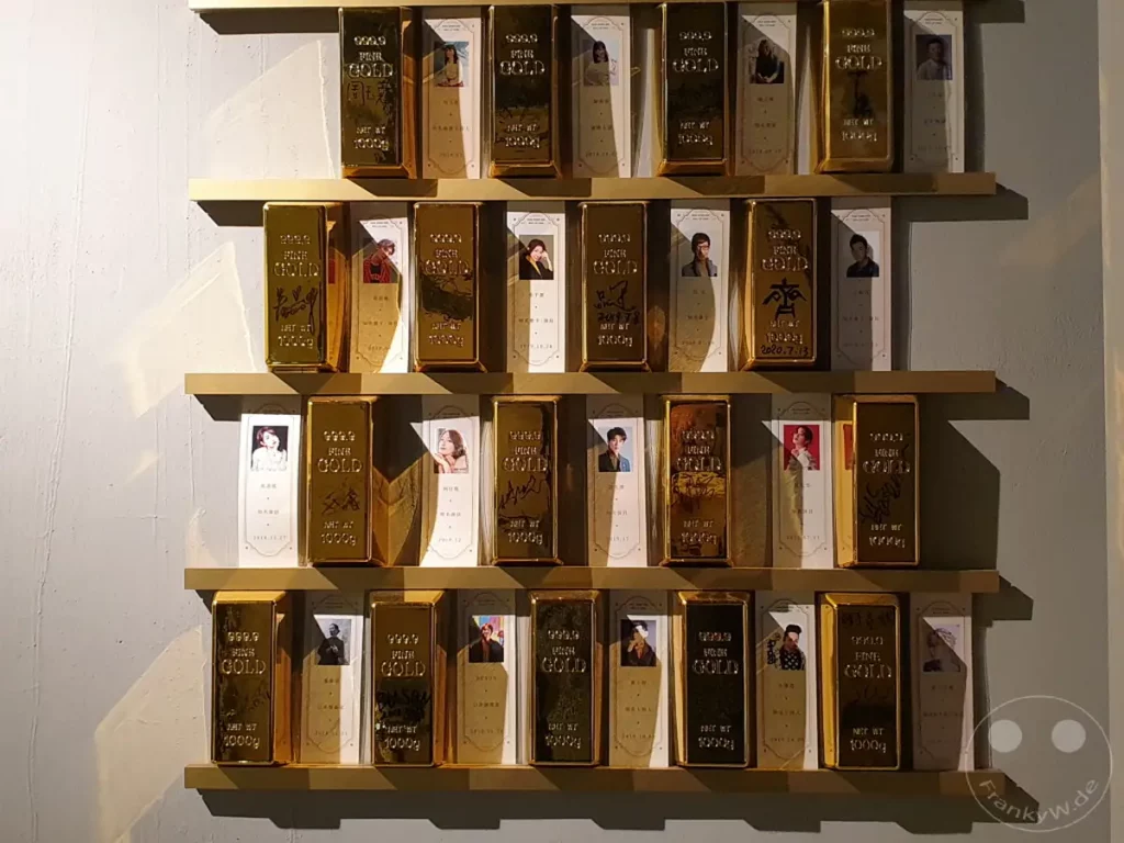 Goldbarren-förmige Souvenirs mit Portraits und Datumsangaben sind ordentlich auf goldenen Regalen in einem Hotel in Taipeh, Taiwan ausgestellt. Die dekorativen Gegenstände erzeugen eine Atmosphäre von Luxus und Erinnerung. Das Arrangement wirkt wie eine Galerie individueller Andenken, die den Aufenthalt in Hotel Hua Shan Din unterstreichen.
