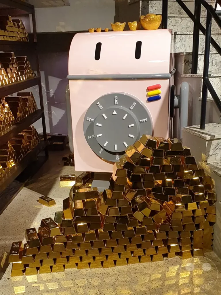 Goldbarren stapeln sich vor einer dekorativen, rosafarbenen Waschmaschine im Hotel Hua Shan Din in Taipeh, Taiwan. Die Szene strahlt eine Atmosphäre von Reichtum und spielerischem Design aus, mit glänzenden Dekorationselementen, die auf dem Gerät und im Umfeld platziert sind. Die Kombination aus einem alltäglichen Haushaltsgerät und einer Fülle von Gold vermittelt eine einzigartige und aufmerksamkeitsstarke visuelle Botschaft.
