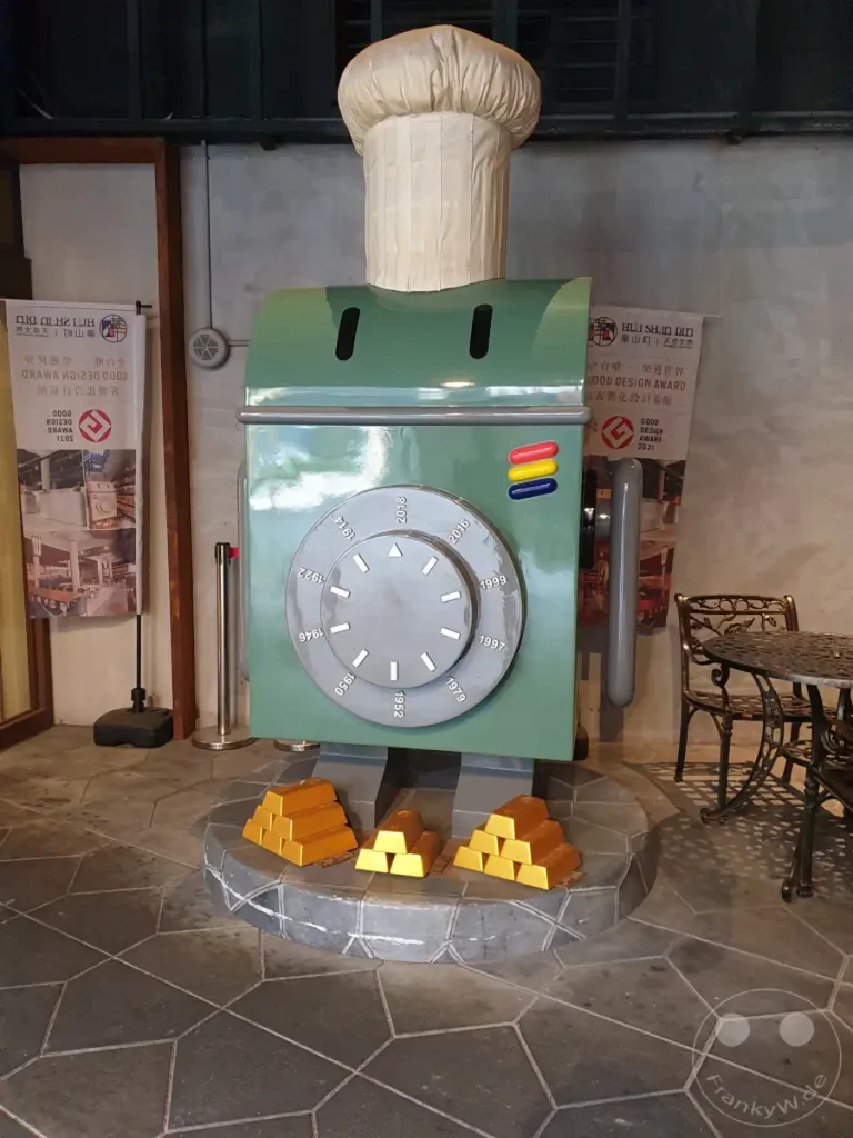 In Taipeh, Taiwan, präsentiert sich das Hotel Hua Shan Din mit einer auffälligen Dekoration, die an einen Kochroboter mit Kochmütze und einer großen Uhr erinnert. Vor der Skulptur sind mehrere gestapelte Goldbarren platziert, die einen Hauch von Opulenz und Einzigartigkeit vermitteln. Die Szene spielt in einem Innenbereich mit industriellem Flair, ergänzt durch ein detailreiches Tischset im Hintergrund.
